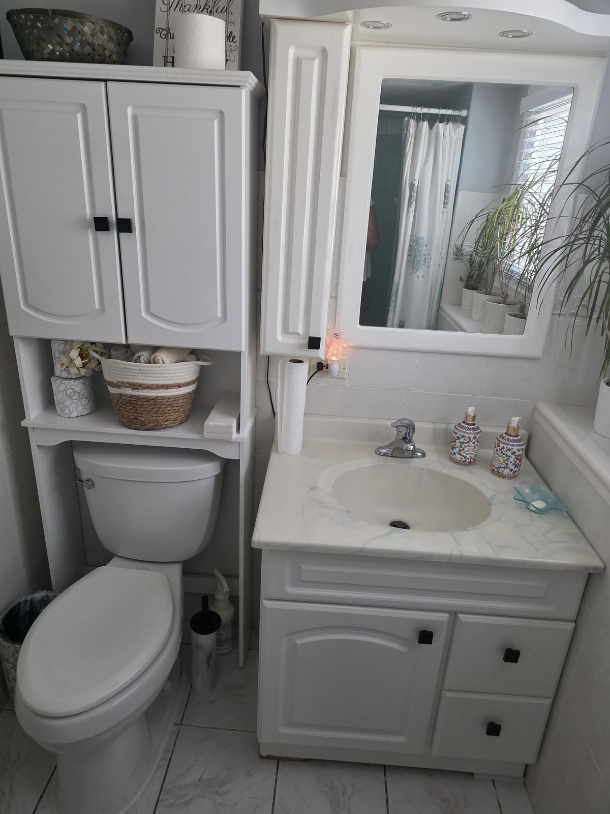 Before: a dated bathroom with a white over-toilet cabinet, a marbled vanity top and a framed mirror